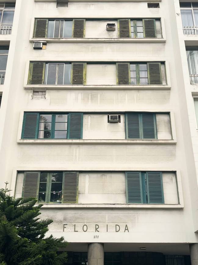 Fachada de um edif&iacute;cio branco com janelas e persianas verdes, algumas abertas, outras fechadas. H&aacute; aparelhos de ar condicionado antigos em algumas janelas. Na parte inferior, o nome FLORIDA e o n&uacute;mero 111 est&atilde;o gravados acima da entrada, com uma &aacute;rvore verde &agrave; esquerda