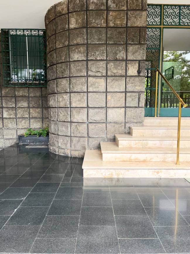 Entrada de um edif&iacute;cio com piso de granito escuro, parede curva de blocos de pedra clara, janela com grade verde e escada de m&aacute;rmore bege com corrim&atilde;o dourado