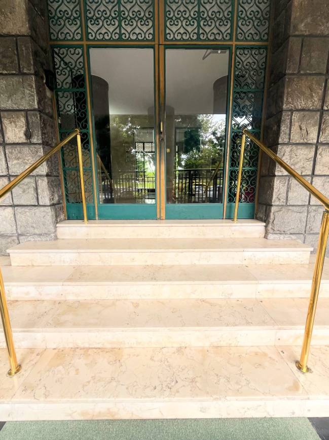 Escadaria de m&aacute;rmore claro com quatro degraus, ladeada por corrim&atilde;os dourados. Ao fundo, uma porta dupla de vidro com detalhes em ferro forjado verde e dourado, refletindo &aacute;rvores e um ambiente externo