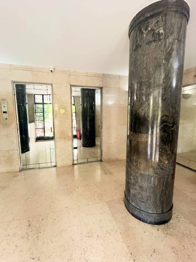 Hall de elevadores com piso e paredes de m&aacute;rmore bege, duas portas de elevador espelhadas e uma coluna cil&iacute;ndrica de m&aacute;rmore escuro &agrave; direita. Uma c&acirc;mera de seguran&ccedil;a est&aacute; no teto