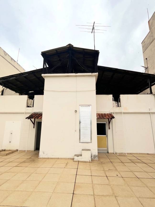 Terra&ccedil;o de um pr&eacute;dio com piso de lajotas bege, paredes brancas e um telhado escuro com antena. H&aacute; uma porta amarela e uma janela vertical de persianas met&aacute;licas, al&eacute;m de duas escadas brancas encostadas nas paredes