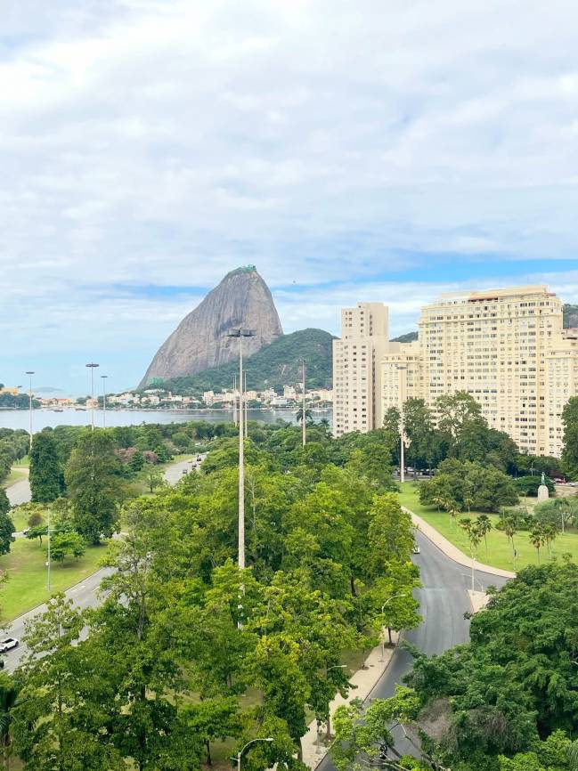 Vista a&eacute;rea do P&atilde;o de A&ccedil;&uacute;car no Rio de Janeiro, com o morro imponente ao fundo. Em primeiro plano, uma &aacute;rea verde com &aacute;rvores frondosas e estradas sinuosas. &Agrave; direita, pr&eacute;dios residenciais altos e brancos. O c&eacute;u est&aacute; parcialmente nublado, com nuvens esparsas e tons de azul claro. A ba&iacute;a se estende &agrave; esquerda, com barcos e a orla da cidade.