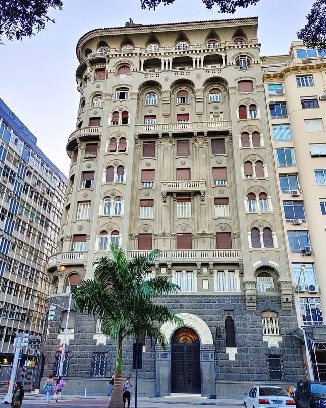 Edif&iacute;cio hist&oacute;rico de fachada cinza com janelas em arco e persianas marrons, varandas ornamentadas e base de pedra escura. Uma palmeira verde est&aacute; em frente, com pessoas caminhando na cal&ccedil;ada e carros estacionados na rua. Outros pr&eacute;dios modernos aparecem ao redor
