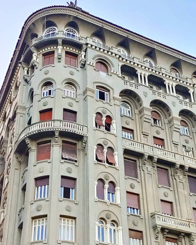 Edif&iacute;cio antigo de esquina com fachada verde-acinzentada, janelas com persianas marrons e detalhes arquitet&ocirc;nicos ornamentados, incluindo arcos, colunas e sacadas com balaustradas brancas, sob um c&eacute;u claro