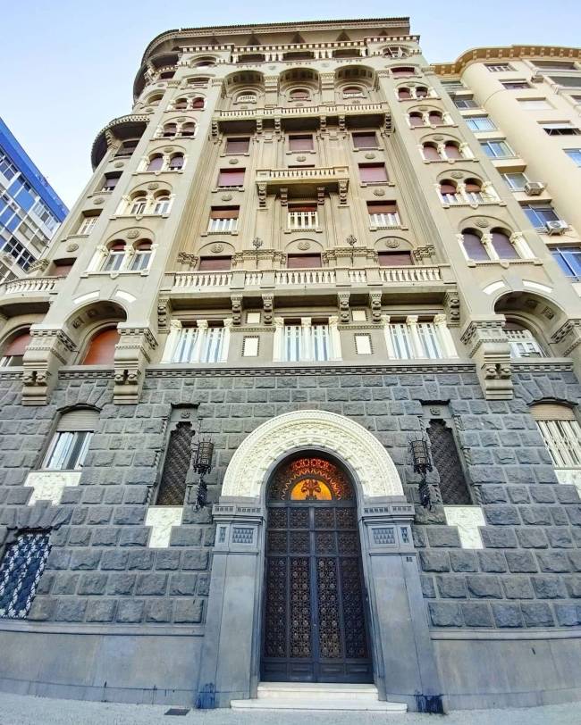 Edif&iacute;cio antigo de estilo ecl&eacute;tico, com fachada cinza-claro e detalhes ornamentados, janelas com persianas vermelhas e sacadas. O t&eacute;rreo possui revestimento de pedras escuras, porta de ferro trabalhada sob um arco branco e vitral colorido no topo