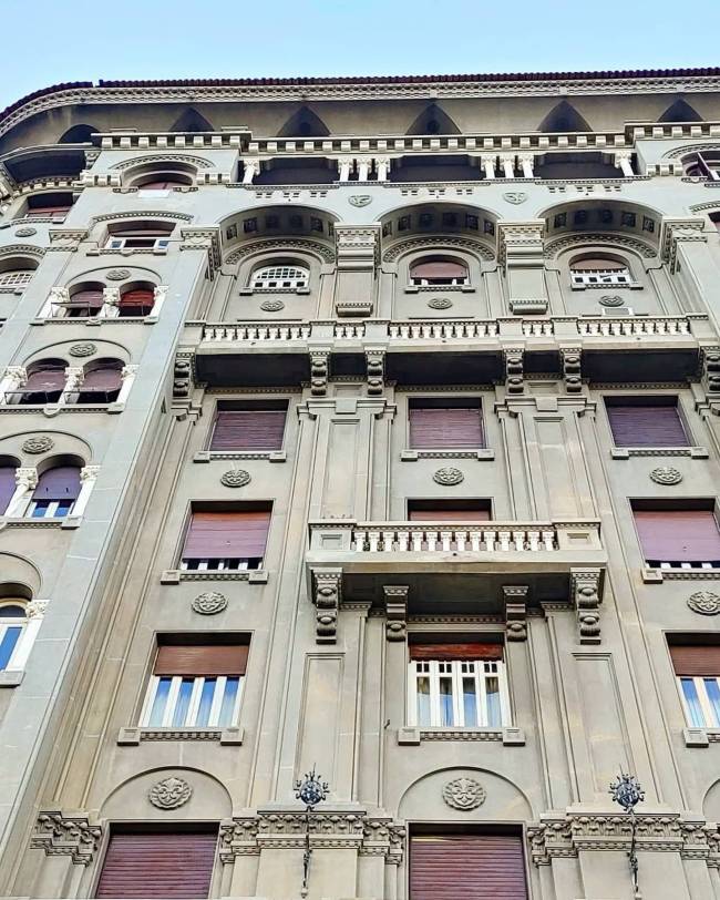 Fachada de um edif&iacute;cio antigo com arquitetura cl&aacute;ssica, detalhes ornamentados, janelas com persianas marrons e um balc&atilde;o central. O pr&eacute;dio &eacute; cinza claro, com molduras e relevos decorativos, visto de baixo para cima contra um c&eacute;u azul
