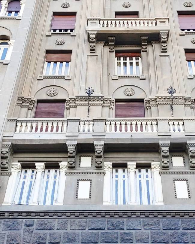 Fachada de edif&iacute;cio antigo com janelas de persianas marrons e sacadas ornamentadas, em tons de cinza e branco, com base de pedras escuras