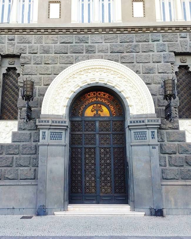 Fachada de edif&iacute;cio com porta dupla de madeira escura e detalhes em ferro forjado, sob arco branco ornamentado. A parede &eacute; de blocos de pedra cinza, com janelas gradeadas e lumin&aacute;rias laterais. Acima, janelas retangulares com molduras brancas e um n&uacute;mero 86 na lateral direita da porta