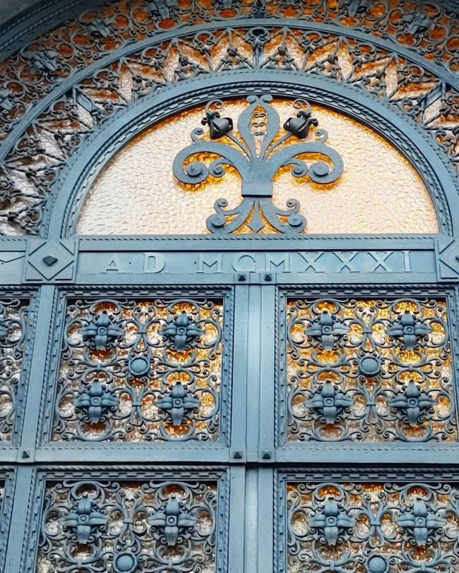 Port&atilde;o de ferro forjado azul-acinzentado com detalhes ornamentados e vidro texturizado &acirc;mbar. Acima, um arco com a inscri&ccedil;&atilde;o A.D. MCMXXI e um bras&atilde;o central em forma de &aacute;rvore estilizada com dois p&aacute;ssaros. Abaixo, pain&eacute;is retangulares com arabescos e flores em relevo, revelando o vidro brilhante por tr&aacute;s