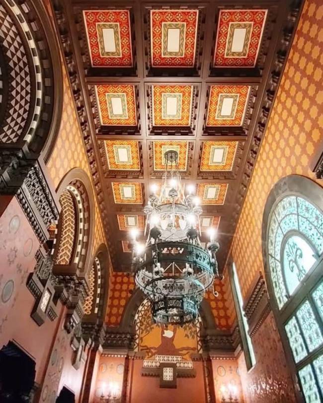 Teto ornamentado com lustres e paredes decoradas em tons terrosos. Um grande lustre de ferro forjado e vidro pende do centro, iluminando o ambiente. O teto possui pain&eacute;is quadrados com padr&otilde;es geom&eacute;tricos em laranja, vermelho e creme. As paredes t&ecirc;m arcos e detalhes em madeira escura, com janelas de vitral &agrave; direita.