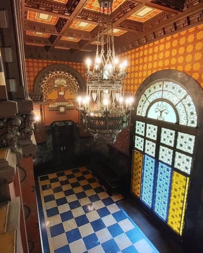 Interior de um sal&atilde;o antigo com teto de madeira entalhada e um grande lustre de ferro forjado com muitas l&acirc;mpadas acesas. As paredes s&atilde;o laranja com padr&otilde;es geom&eacute;tricos. No ch&atilde;o, um piso xadrez azul e branco. &Agrave; direita, uma porta alta com vitrais coloridos em tons de azul e amarelo, e acima, uma janela semicircular com vitral branco. Ao fundo, um arco com um mural de &aacute;rvore. A cena &eacute; vista de cima, de uma escadaria
