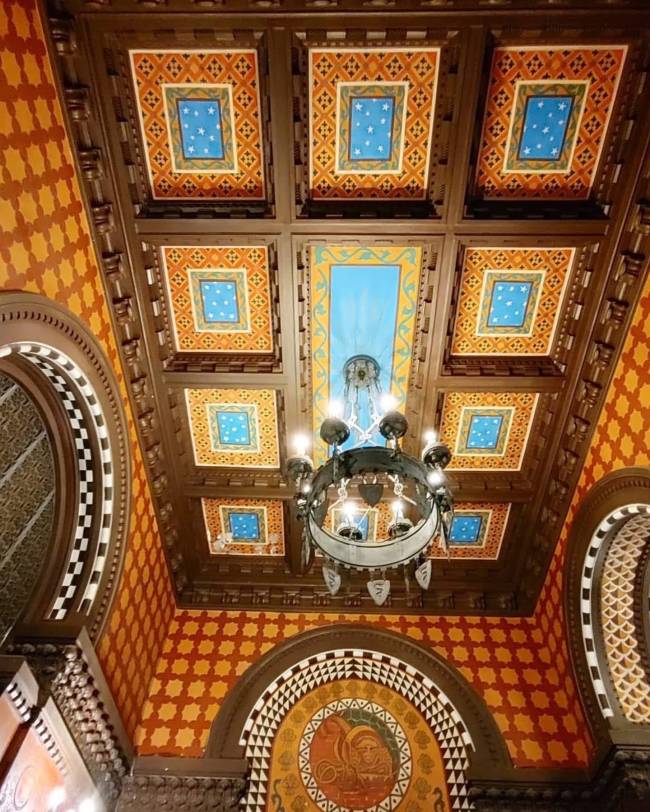 Teto e paredes ricamente decorados com padr&otilde;es geom&eacute;tricos e cores vibrantes. O teto possui caixot&otilde;es de madeira escura, com pain&eacute;is quadrados em tons de laranja, azul e amarelo, alguns com estrelas. Um grande lustre de metal escuro com v&aacute;rias l&acirc;mpadas pendura no centro. As paredes exibem um padr&atilde;o laranja e marrom, com arcos decorados em preto, branco e tons terrosos, apresentando figuras estilizadas