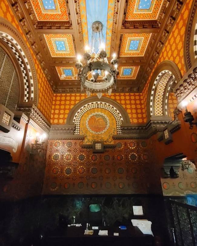 Interior de um sal&atilde;o com teto e paredes ricamente decorados em estilo mourisco. O teto tem pain&eacute;is quadrados em tons de laranja, azul e madeira escura, com um grande lustre de ferro forjado no centro. As paredes exibem padr&otilde;es geom&eacute;tricos repetitivos em tons terrosos e um arco com mosaico circular. Uma bancada escura ocupa a parte inferior da imagem, com alguns objetos sobre ela