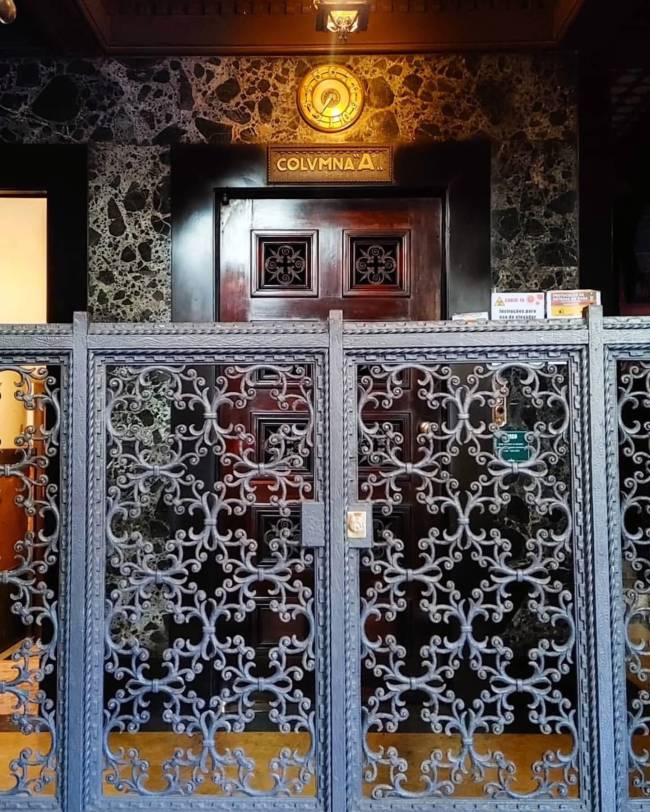 Port&atilde;o de ferro cinza com arabescos intrincados, revelando uma porta de madeira escura com detalhes entalhados. Acima, uma placa dourada com COLUMNA A e um rel&oacute;gio redondo, tudo em uma parede de m&aacute;rmore escuro com veios claros