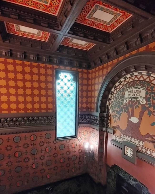 Interior de um edif&iacute;cio com teto de madeira escura e pain&eacute;is decorados em vermelho e laranja. As paredes t&ecirc;m padr&otilde;es geom&eacute;tricos em tons de laranja e vermelho, com um vitral azul claro vertical. &Agrave; direita, um arco revela um mural com uma &aacute;rvore e a inscri&ccedil;&atilde;o ANNO DOMINI MCMXXXI