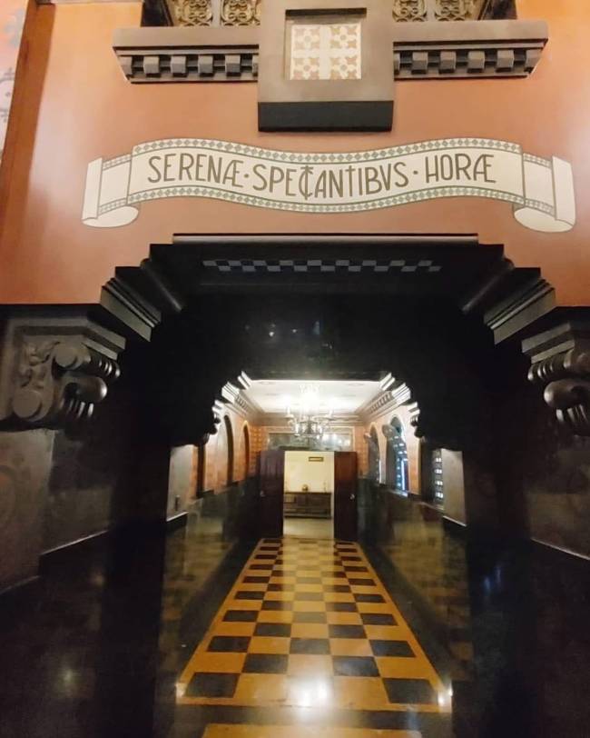 Corredor de um edif&iacute;cio antigo com piso xadrez preto e amarelo, paredes escuras e um arco decorado. Acima do arco, uma faixa branca com a inscri&ccedil;&atilde;o em latim SERENAE SPECANTIBUS HORAE em letras escuras, sobre uma parede cor de salm&atilde;o. No fundo, um ambiente iluminado com um lustre.