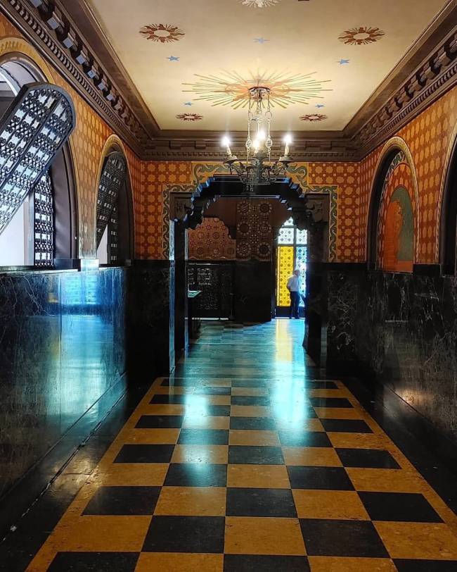 Corredor longo com piso xadrez preto e amarelo, paredes em tons de laranja e marrom com padr&otilde;es geom&eacute;tricos. O teto bege tem um lustre central e decora&ccedil;&otilde;es de estrelas. Arcos ornamentados e uma pessoa ao fundo, em uma porta com vitral colorido, completam a cena