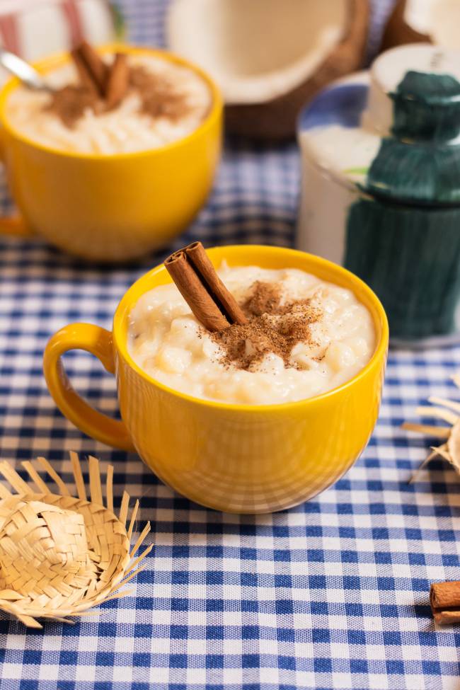 Duas canecas amarelas com arroz doce cremoso, polvilhado com canela em p&oacute; e dois paus de canela, sobre uma toalha xadrez azul e branca. Ao fundo, coco partido e um saleiro. &Agrave; frente, um chap&eacute;u de palha miniatura