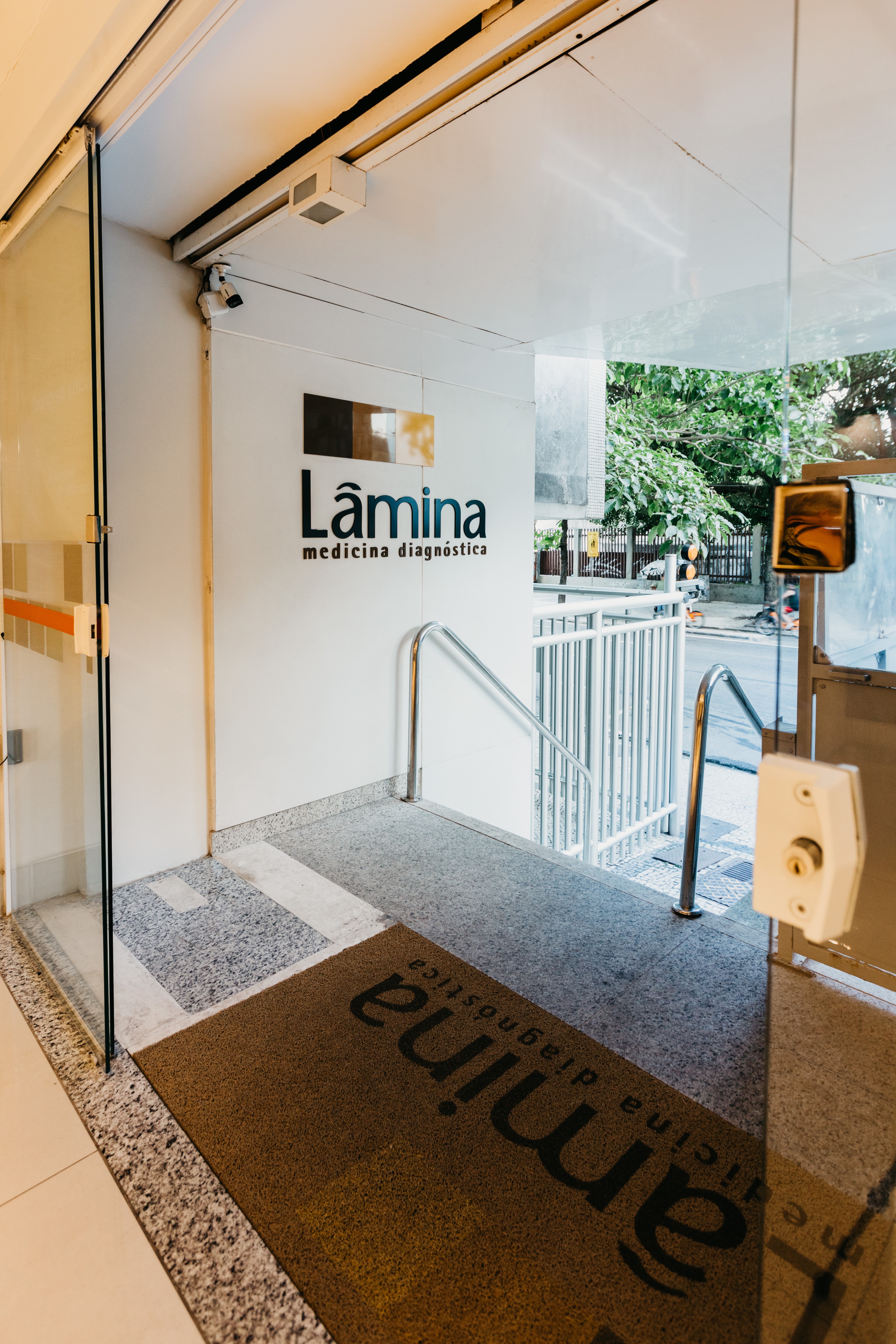 Entrada de uma cl&iacute;nica m&eacute;dica com porta de vidro, tapete marrom escrito L&acirc;mina medicina diagn&oacute;stica e rampa de acesso com corrim&atilde;o de metal. Ao fundo, &aacute;rvores e a rua.