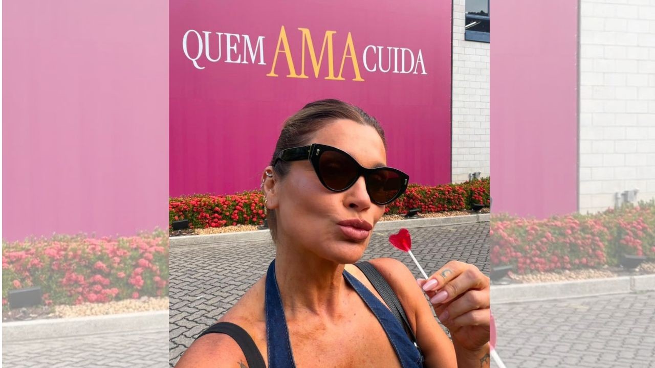 Mulher de óculos escuros e blusa azul, com cabelo preso, faz bico segurando um pirulito de coração vermelho. Ao fundo, um muro rosa com a frase QUEM AMA CUIDA em letras brancas e amarelas, e arbustos floridos