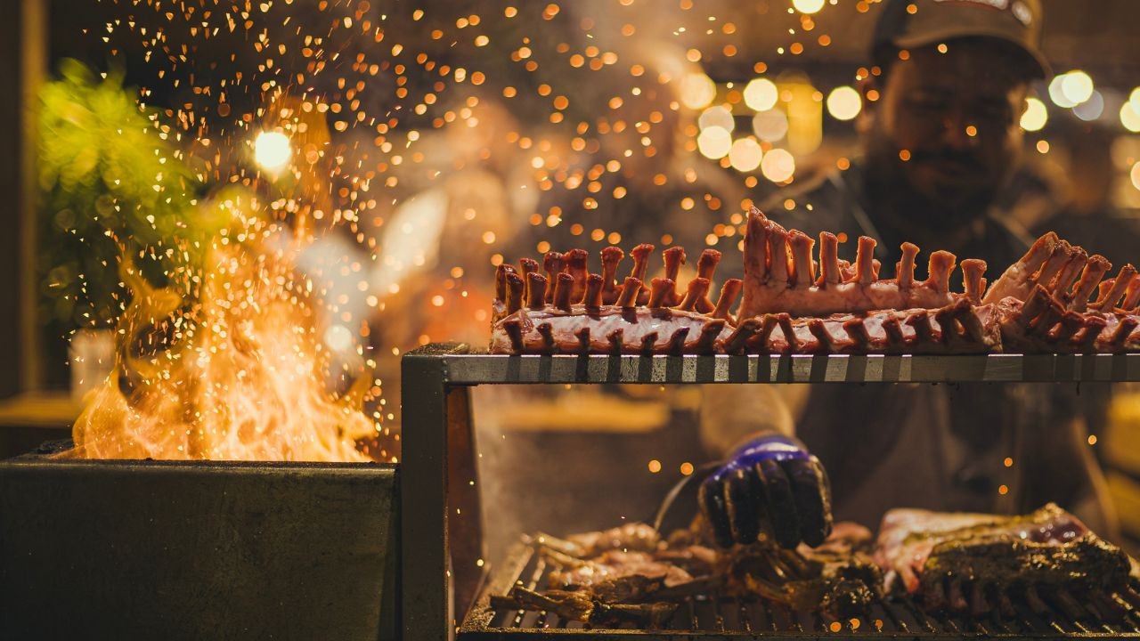 Costelas de carne cruas e temperadas em uma grelha de dois andares, com chamas e faíscas à esquerda. Um homem de boné e luva azul manuseia a carne no fundo, com luzes bokeh douradas desfocadas.