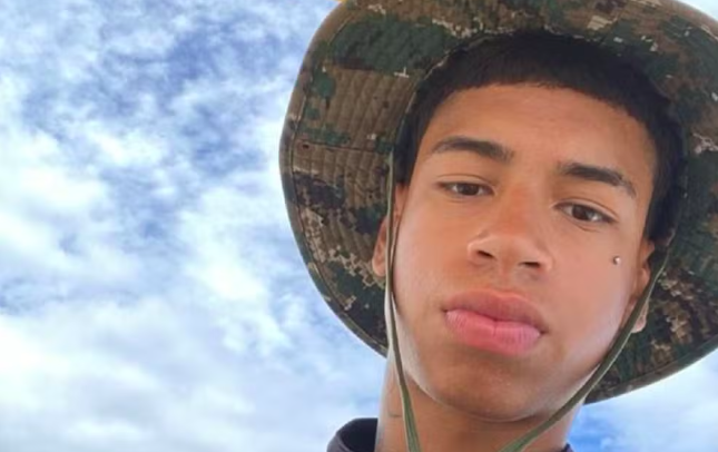 Jovem de pele morena clara, usando chapéu camuflado, com piercing na bochecha esquerda e lábios levemente inchados, olhando para a câmera sob céu azul com nuvens brancas