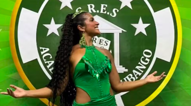 Mulher sorridente de perfil, com cabelo crespo preso em rabo de cavalo alto, usando um vestido verde brilhante com recortes e brincos longos. Ela está com os braços abertos, em frente a um escudo verde com estrelas e o nome G.R.E.S. ACADÊMICOS DO BANGU