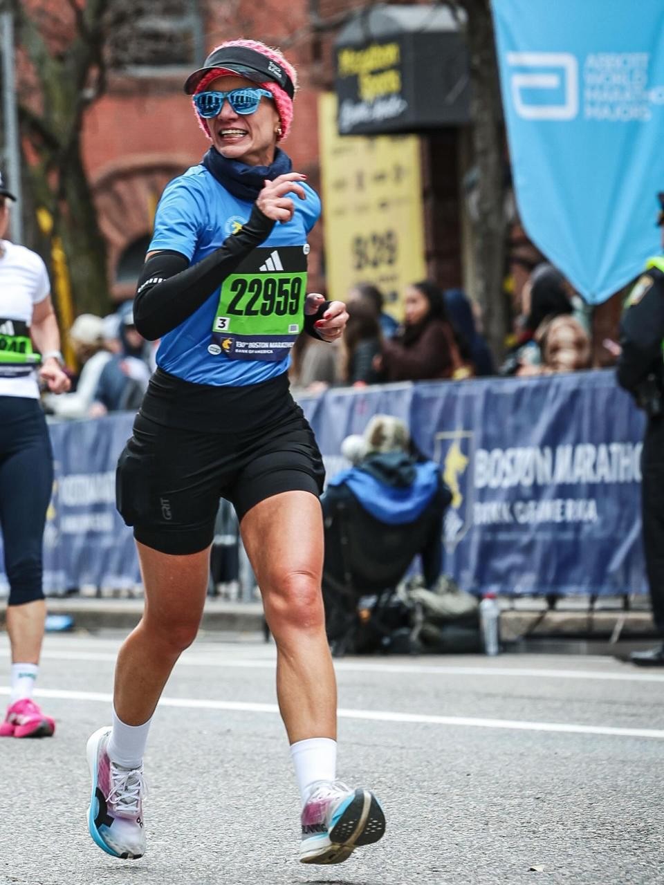 Corredora sorridente, de &oacute;culos de sol azuis e gorro rosa, usando camiseta azul, manguitos e shorts pretos, com o n&uacute;mero 22959 no peito, correndo em uma maratona urbana, com p&uacute;blico e banners ao fundo