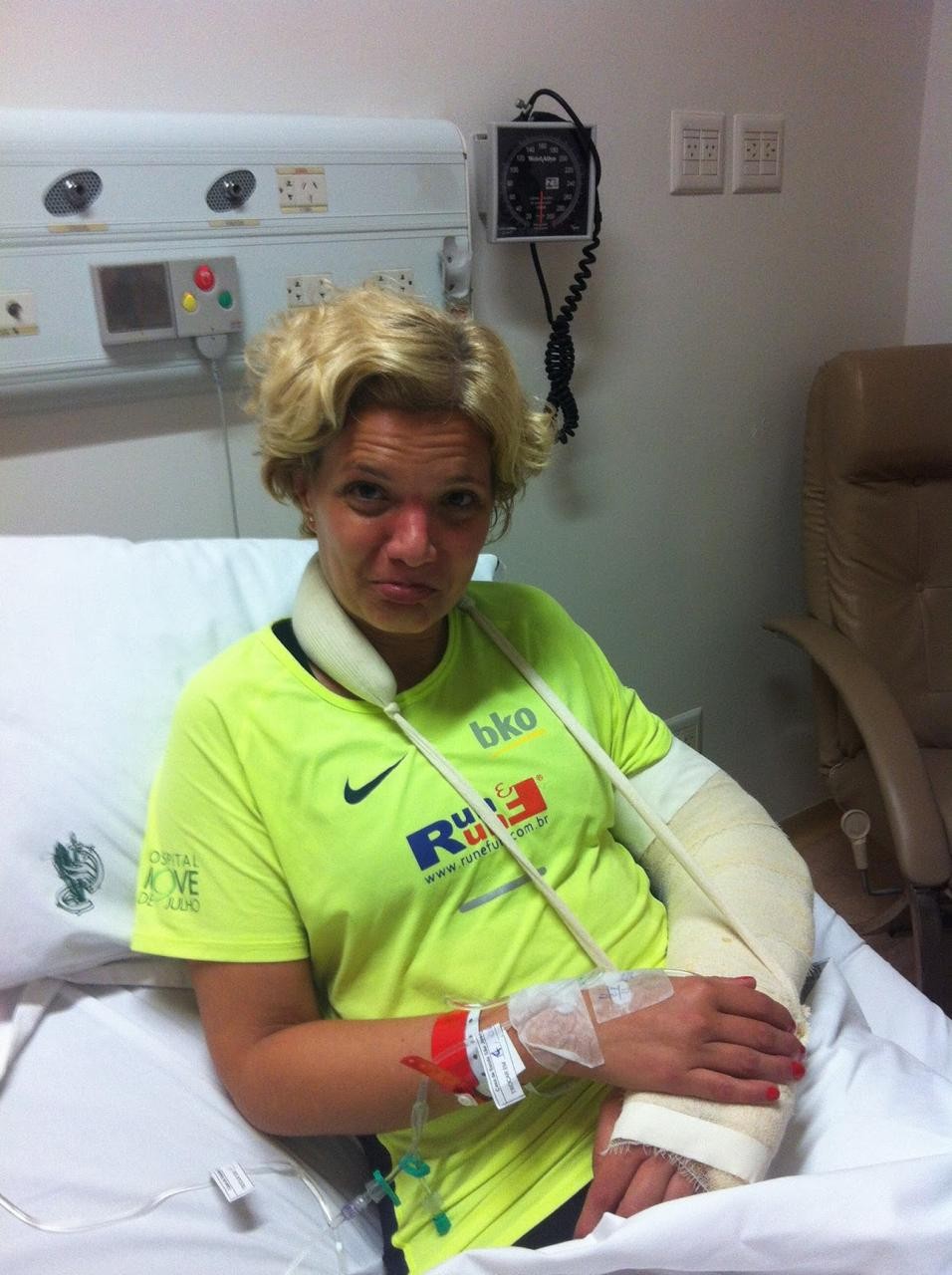 Mulher loira, com express&atilde;o de cansa&ccedil;o, sentada em leito hospitalar, usando tipoia e gesso no bra&ccedil;o esquerdo, com acesso venoso no bra&ccedil;o direito. Veste camiseta verde-lim&atilde;o com logotipos de marcas esportivas. Ao fundo, painel de equipamentos m&eacute;dicos e poltrona marrom