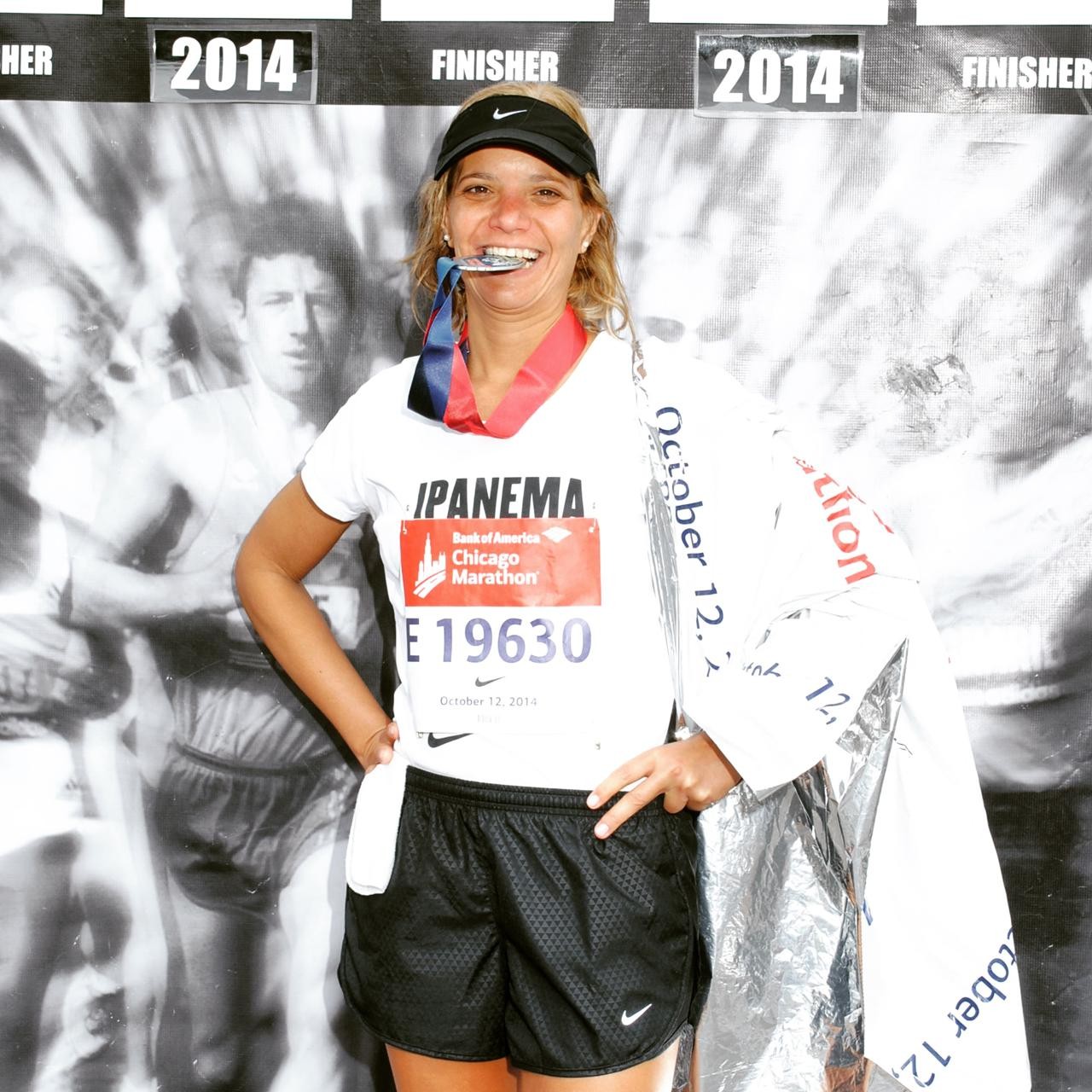 Mulher sorridente com viseira preta e medalha no pesco&ccedil;o, mordendo a medalha. Veste camiseta branca com n&uacute;mero de peito E19630 e shorts pretos, segurando um cobertor t&eacute;rmico prateado. Ao fundo, uma imagem em preto e branco de corredores e a inscri&ccedil;&atilde;o 