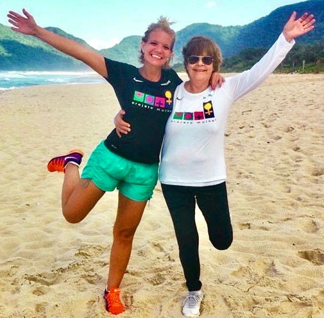 Duas mulheres sorridentes, uma jovem e uma idosa, abra&ccedil;adas na praia. A jovem veste camiseta preta e shorts verde, levantando uma perna. A idosa, de &oacute;culos escuros, usa camiseta branca de manga longa e cal&ccedil;a preta. Ambas exibem camisetas com a estampa projeto mulher e s&iacute;mbolos femininos coloridos. Ao fundo, o mar e montanhas verdes sob um c&eacute;u azul