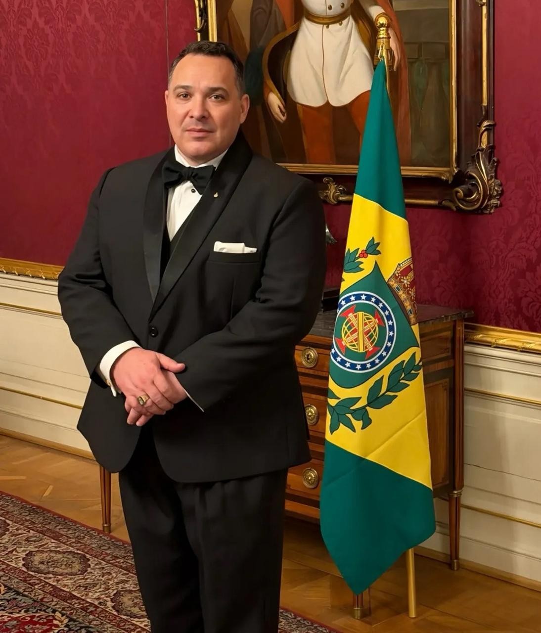 Homem de meia-idade, cabelo escuro, terno preto com gravata borboleta, em p&eacute;, m&atilde;os cruzadas, anel dourado no dedo anelar. Ao lado, uma bandeira verde e amarela com bras&atilde;o. Ao fundo, parede vermelha, quadro e m&oacute;vel de madeira.