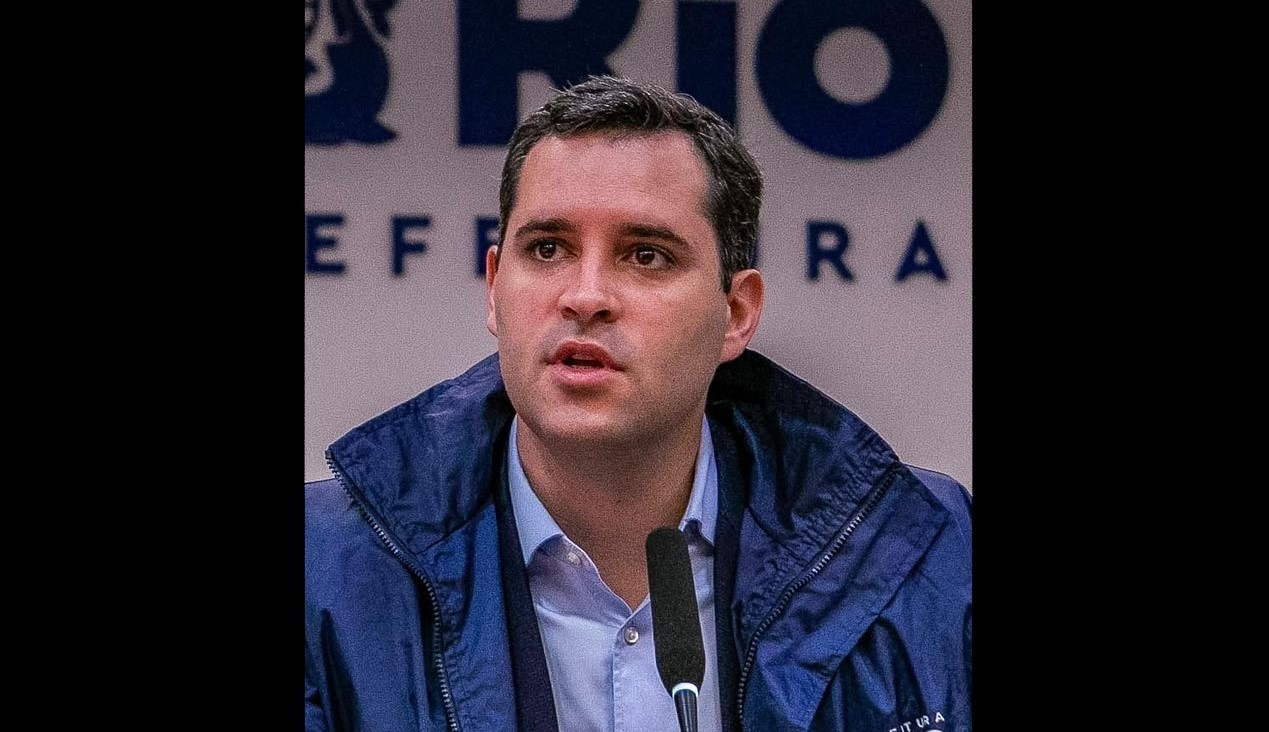 Homem branco de cabelo escuro, vestindo camisa azul clara e jaqueta azul escura, fala em um microfone. Ao fundo, letras brancas "RIO" e "EFE" em um painel azul