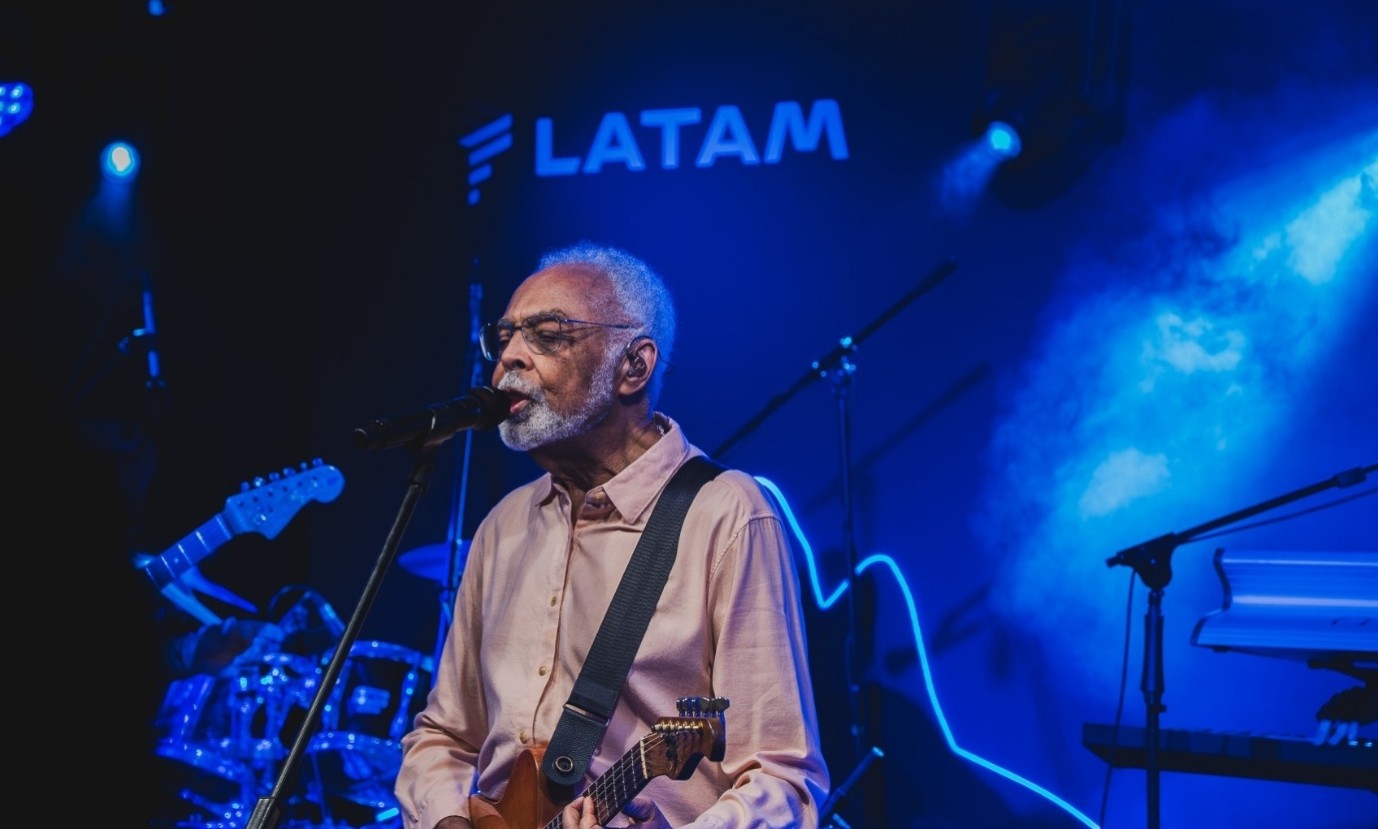 Gilberto Gil, cantor e compositor, com barba e cabelos brancos, óculos, camisa salmão, tocando violão e cantando em microfone. Ele está em um palco com iluminação azul e o logo da LATAM ao fundo