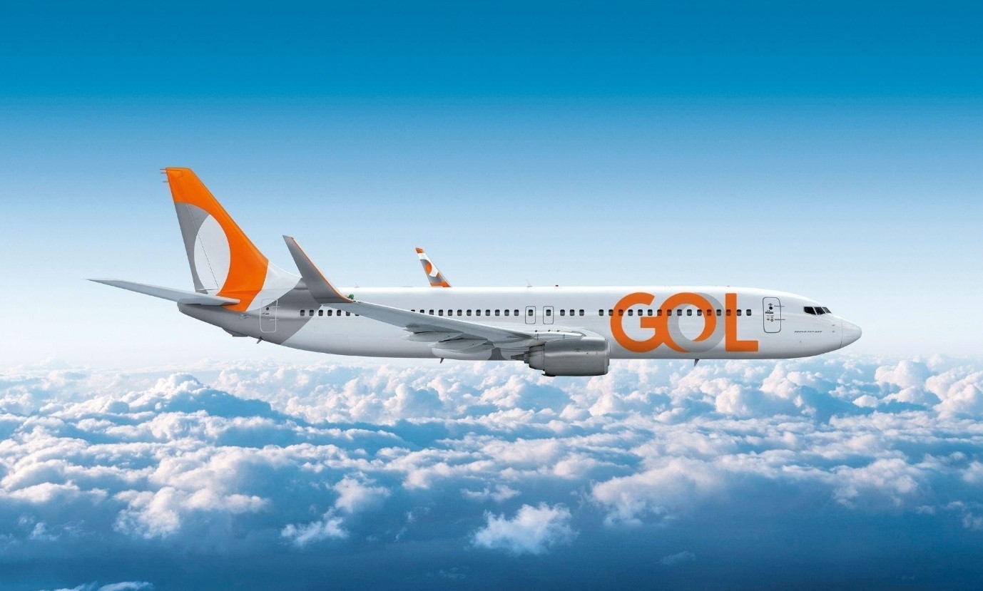 Avião branco da companhia aérea Gol, com a cauda laranja e cinza, e o nome GOL em laranja na fuselagem, voando acima de nuvens brancas sob um céu azul claro