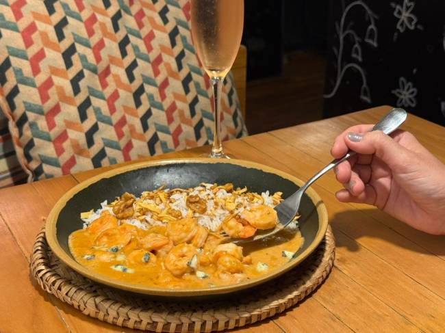 Uma m&atilde;o feminina com unhas cinzas segura um garfo com camar&otilde;es e molho cremoso, sobre um prato de arroz com nozes e cebola frita em uma mesa de madeira. Ao fundo, um copo de champanhe e uma almofada geom&eacute;trica colorida