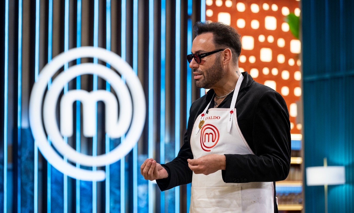 Homem de barba, óculos escuros e cabelo escuro, vestindo avental branco com o logo MasterChef e blazer preto, gesticula com as mãos em um estúdio com fundo azul e o logo MasterChef em destaque