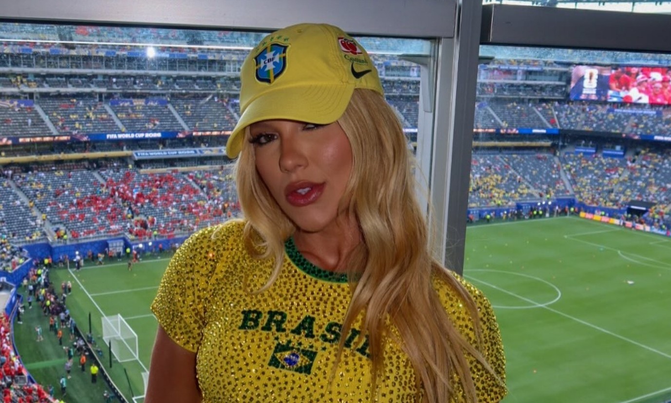 Mulher loira com boné e blusa amarela do Brasil, com brilhos, posa em camarote de estádio de futebol lotado