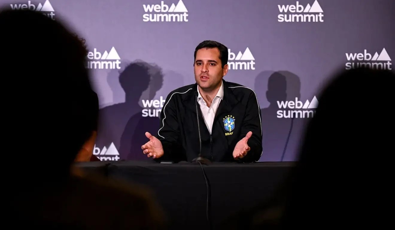 Homem branco de camisa branca e jaqueta preta com emblema do Brasil, gesticulando com as mãos abertas, sentado em uma mesa preta. Ao fundo, uma parede roxa com o logo web summit repetido em branco. Uma silhueta escura de uma pessoa aparece em primeiro plano, à direita