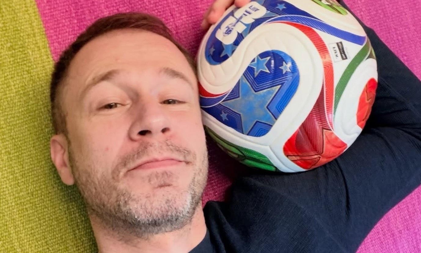 Homem branco de barba rala e cabelo curto castanho claro, sorrindo levemente, deitado de lado com uma bola de futebol colorida encostada na cabeça, sobre um tecido listrado verde e rosa