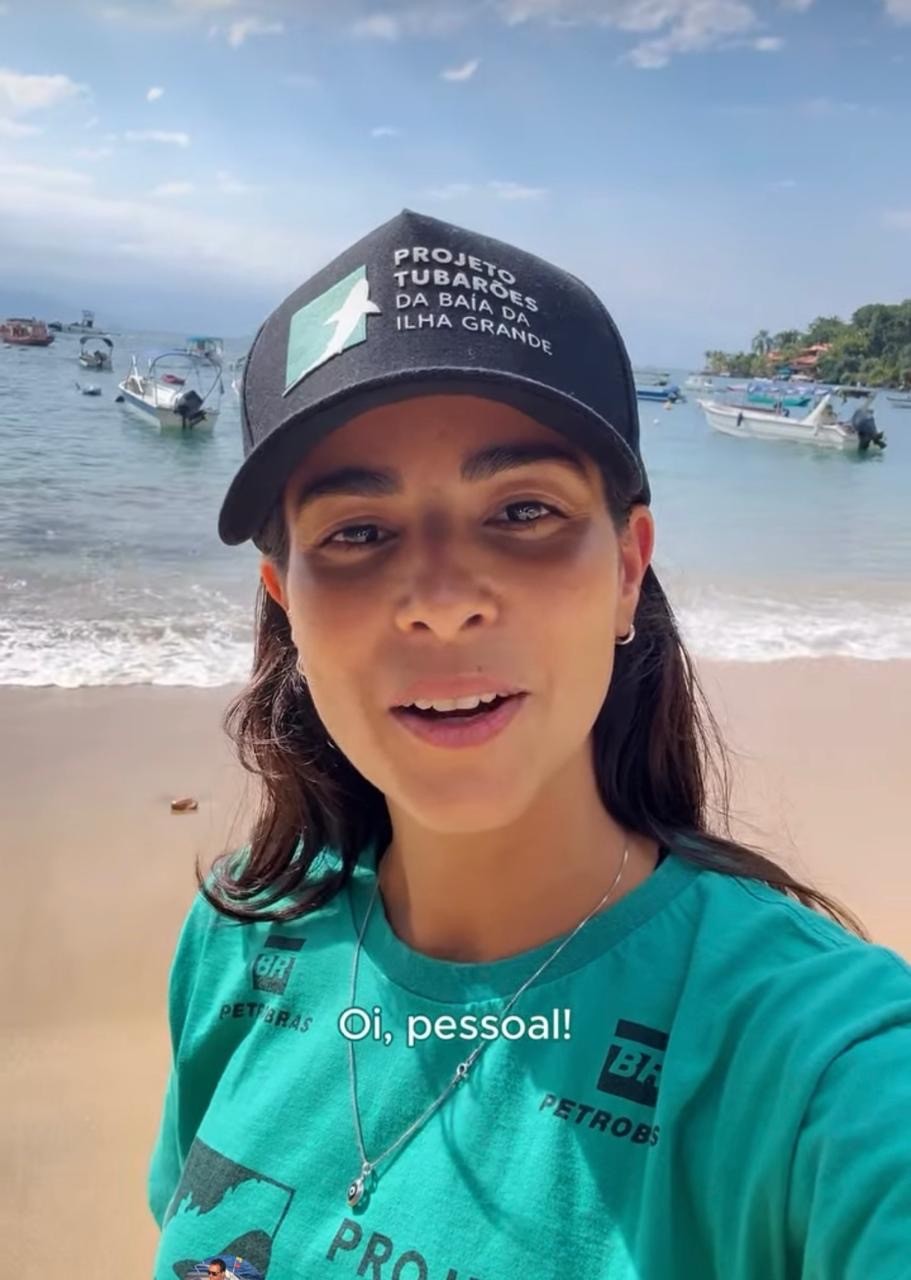 Mulher sorridente, de cabelos escuros, usando bon&eacute; preto com PROJETO TUBAR&Otilde;ES DA BA&Iacute;A DA ILHA GRANDE e camiseta verde com BR PETROBRAS, em uma praia com barcos no mar e vegeta&ccedil;&atilde;o ao fundo