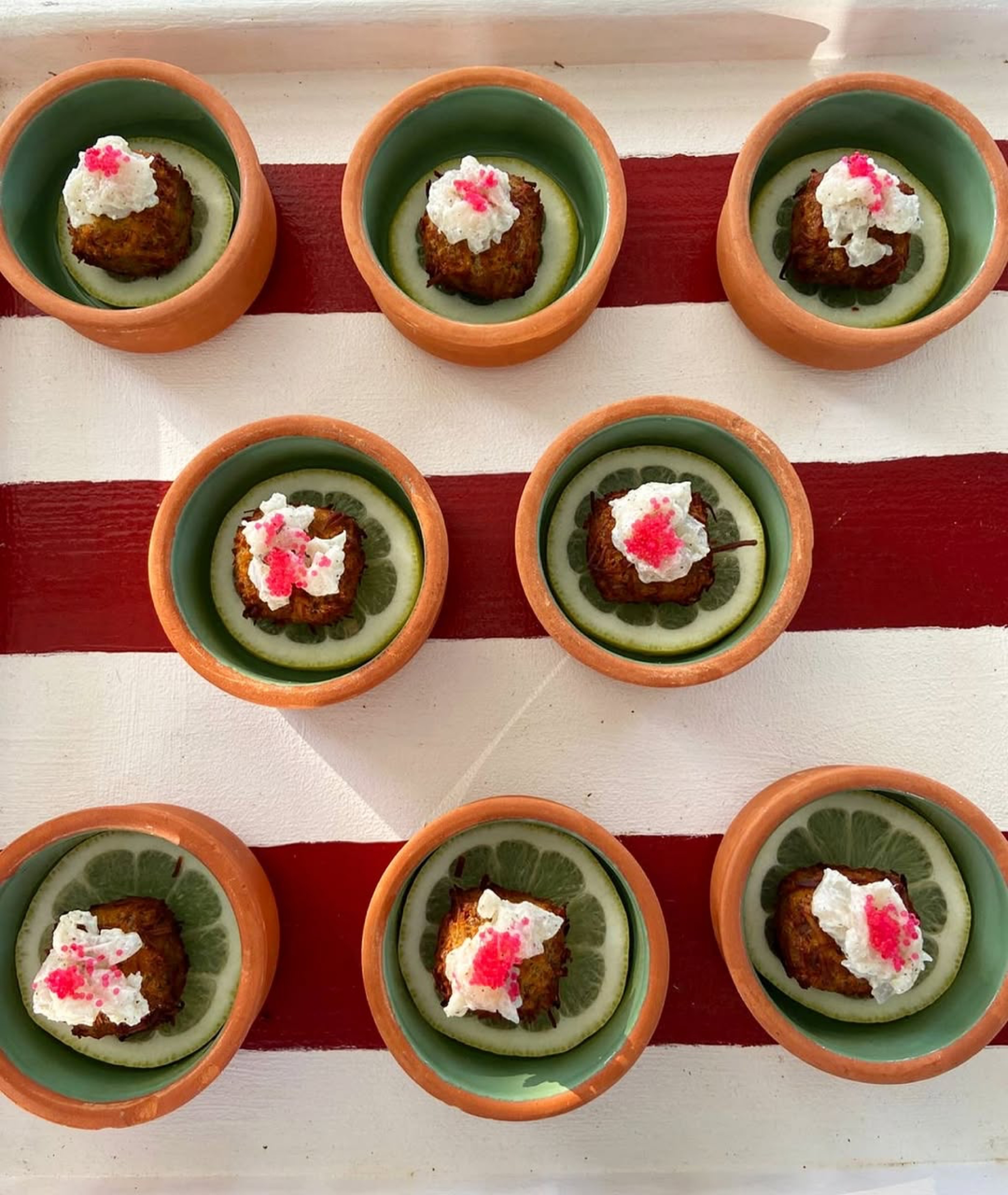 Nove por&ccedil;&otilde;es individuais de bolinhos fritos, cada um sobre uma rodela de lim&atilde;o, servidos em potes de barro com interior verde, dispostos em um tabuleiro branco com listras vermelhas. Os bolinhos s&atilde;o cobertos com um creme branco e granulado rosa