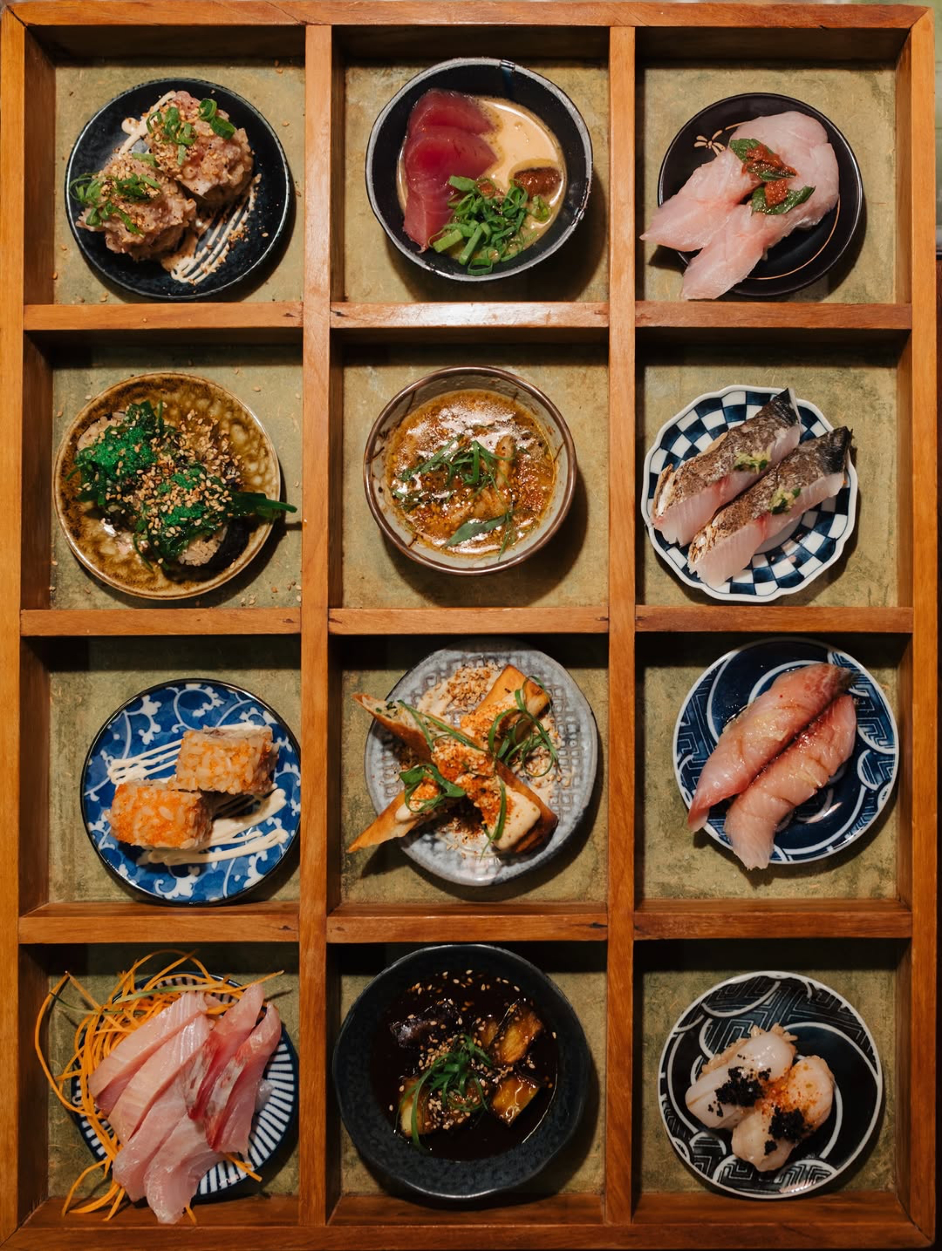 Nove pratos de comida japonesa em nichos de madeira, incluindo sushis, sashimis, temakis e outros petiscos com peixes, vegetais e molhos variados