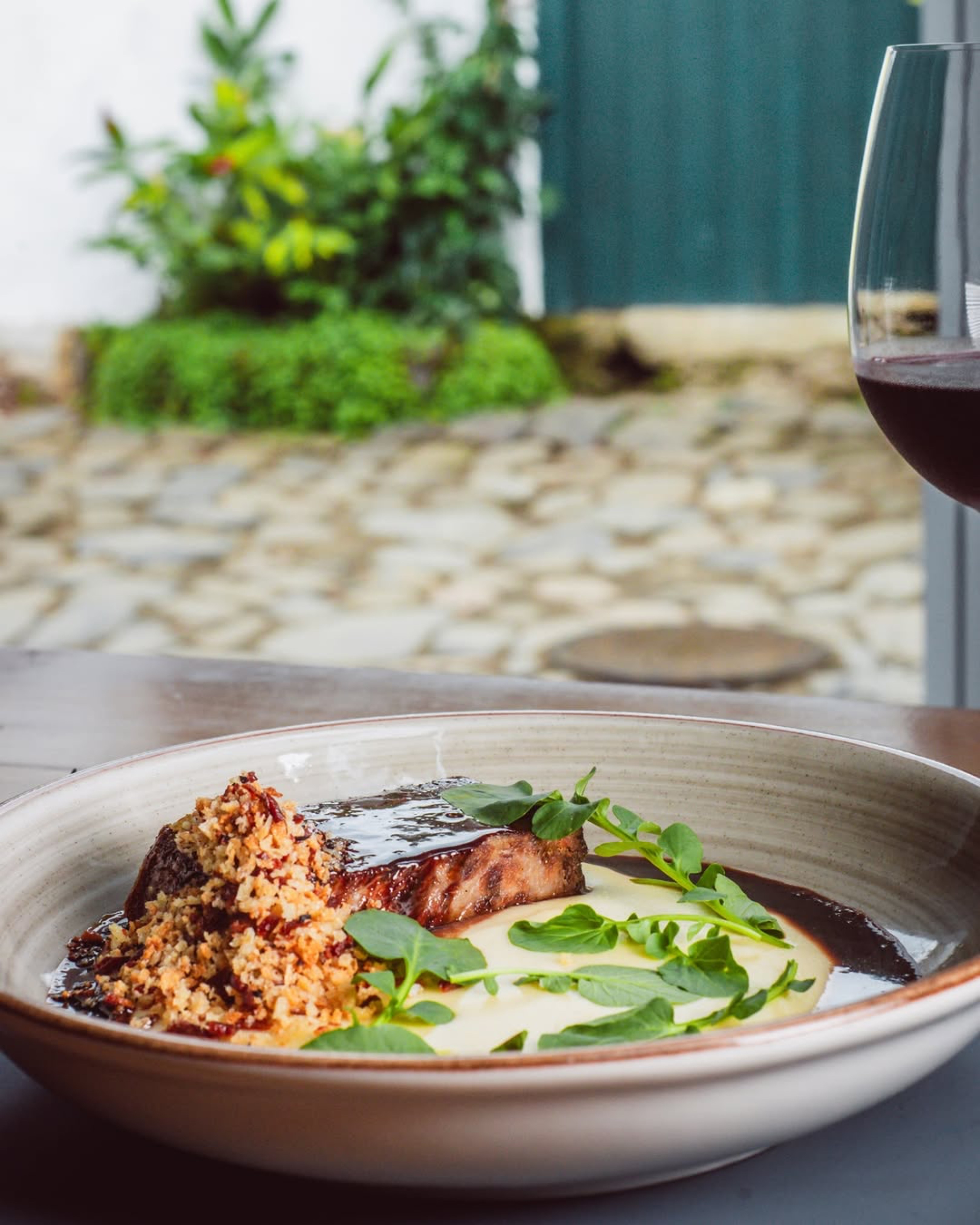 Prato gourmet com carne grelhada coberta por molho escuro, pur&ecirc; cremoso, farofa crocante e folhas verdes, ao lado de uma ta&ccedil;a de vinho tinto, com fundo desfocado de um p&aacute;tio de pedras e plantas