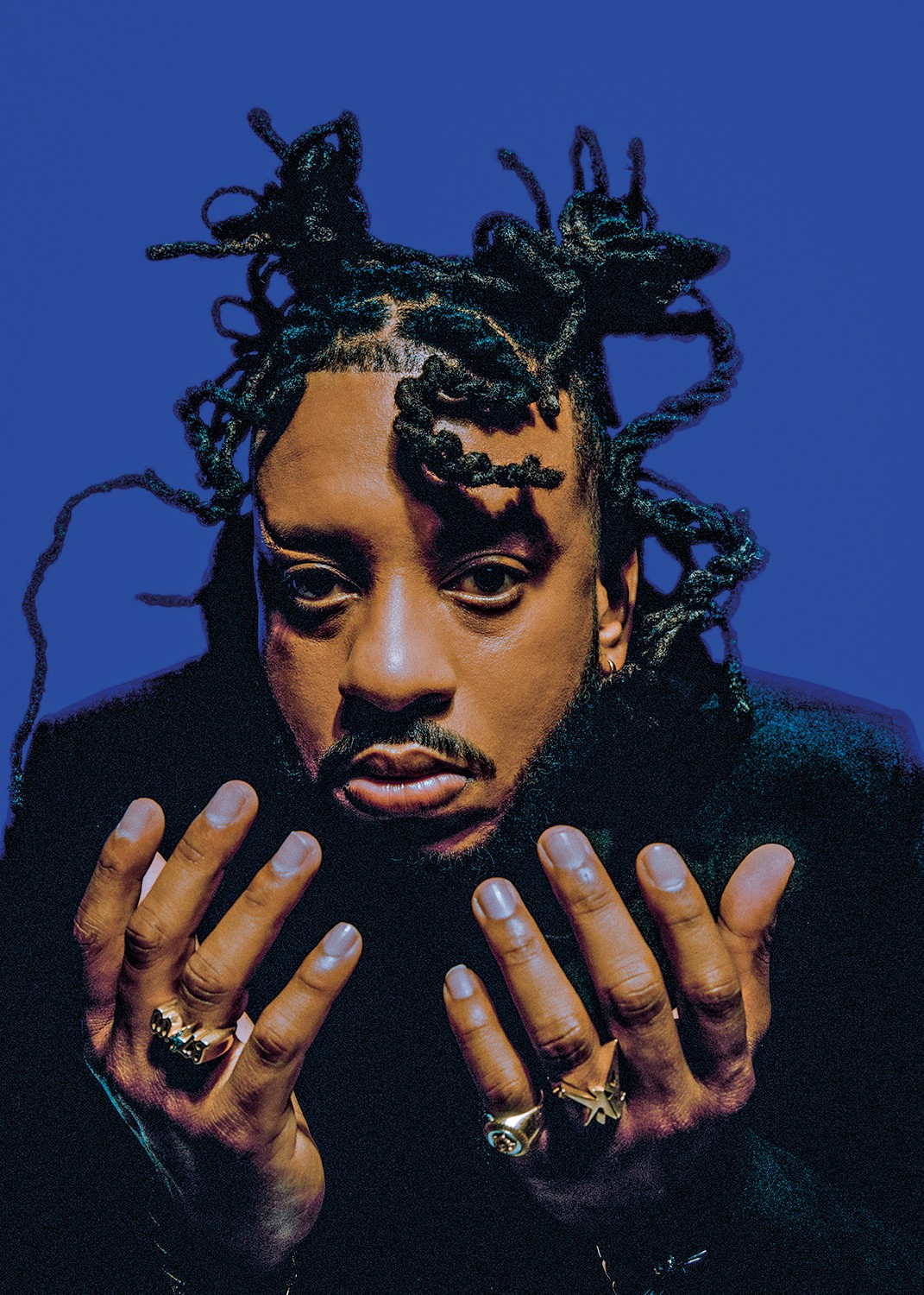 Homem negro com dreads presos para cima, barba e bigode, veste casaco escuro e olha fixamente. Ele exibe an&eacute;is dourados nas m&atilde;os abertas, em fundo azul