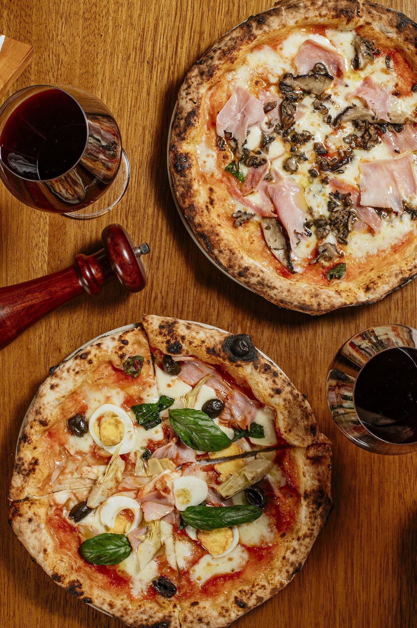 Duas pizzas redondas com bordas crocantes em uma mesa de madeira. A superior tem presunto, cogumelos e manjeric&atilde;o. A inferior, fatiada, tem presunto, ovos de codorna, azeitonas pretas, alcachofras e manjeric&atilde;o. Dois copos de vinho tinto e um moedor de pimenta vermelho completam a cena.