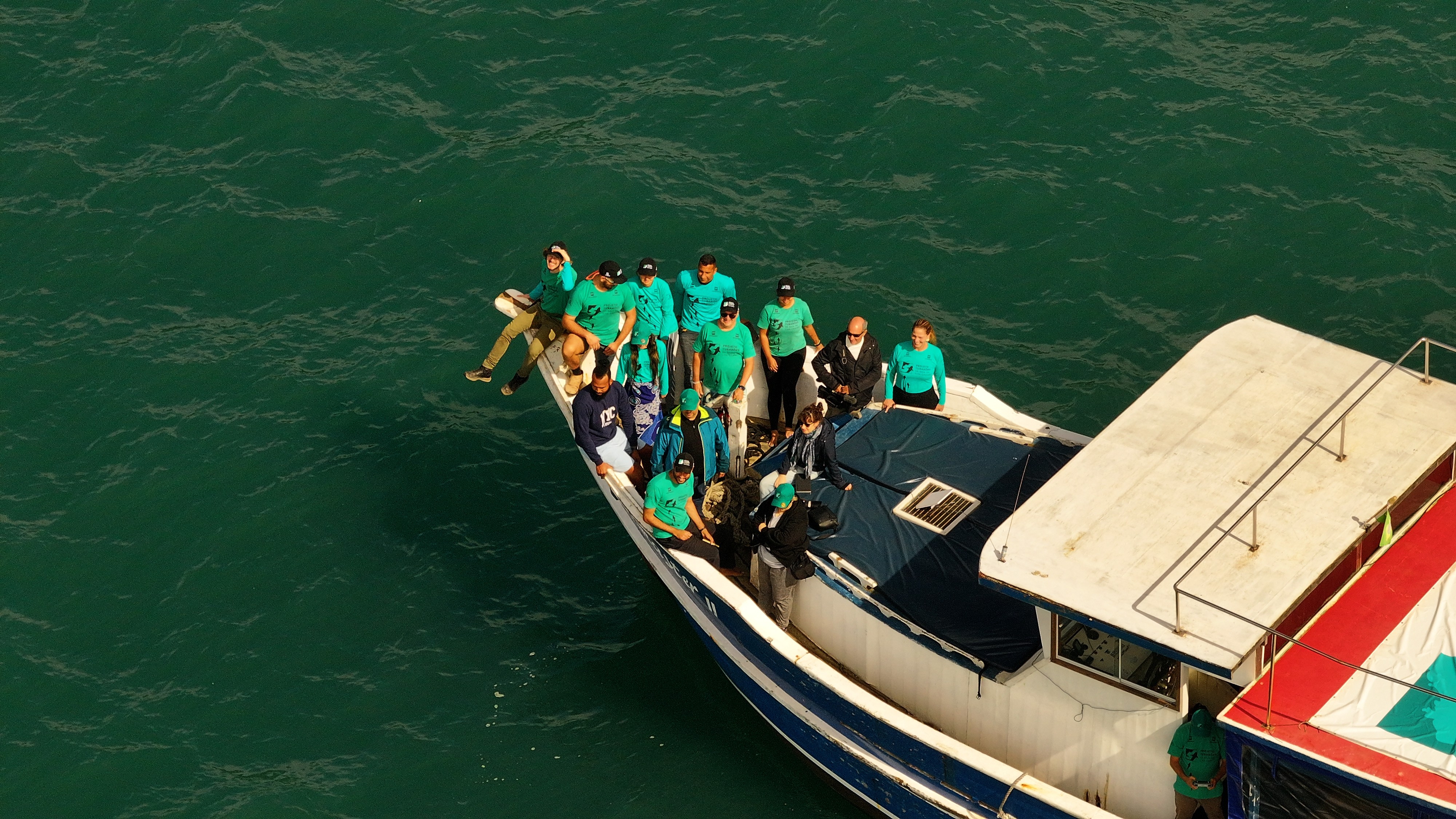 Vista a&eacute;rea de um grupo de pessoas em um barco branco e azul, navegando em &aacute;guas verde-escuras. A maioria veste camisetas verde-&aacute;gua, alguns est&atilde;o na proa, outros no conv&eacute;s. Uma pessoa senta na borda da proa, com as pernas para fora.