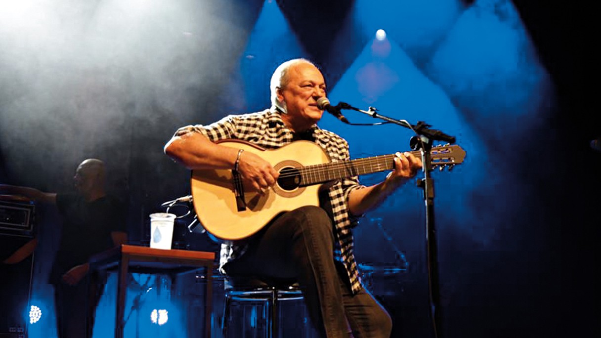 Homem de cabelos brancos, camisa xadrez e cal&ccedil;a escura, sentado, tocando viol&atilde;o e cantando em microfone, sob luzes azuis e fuma&ccedil;a no palco