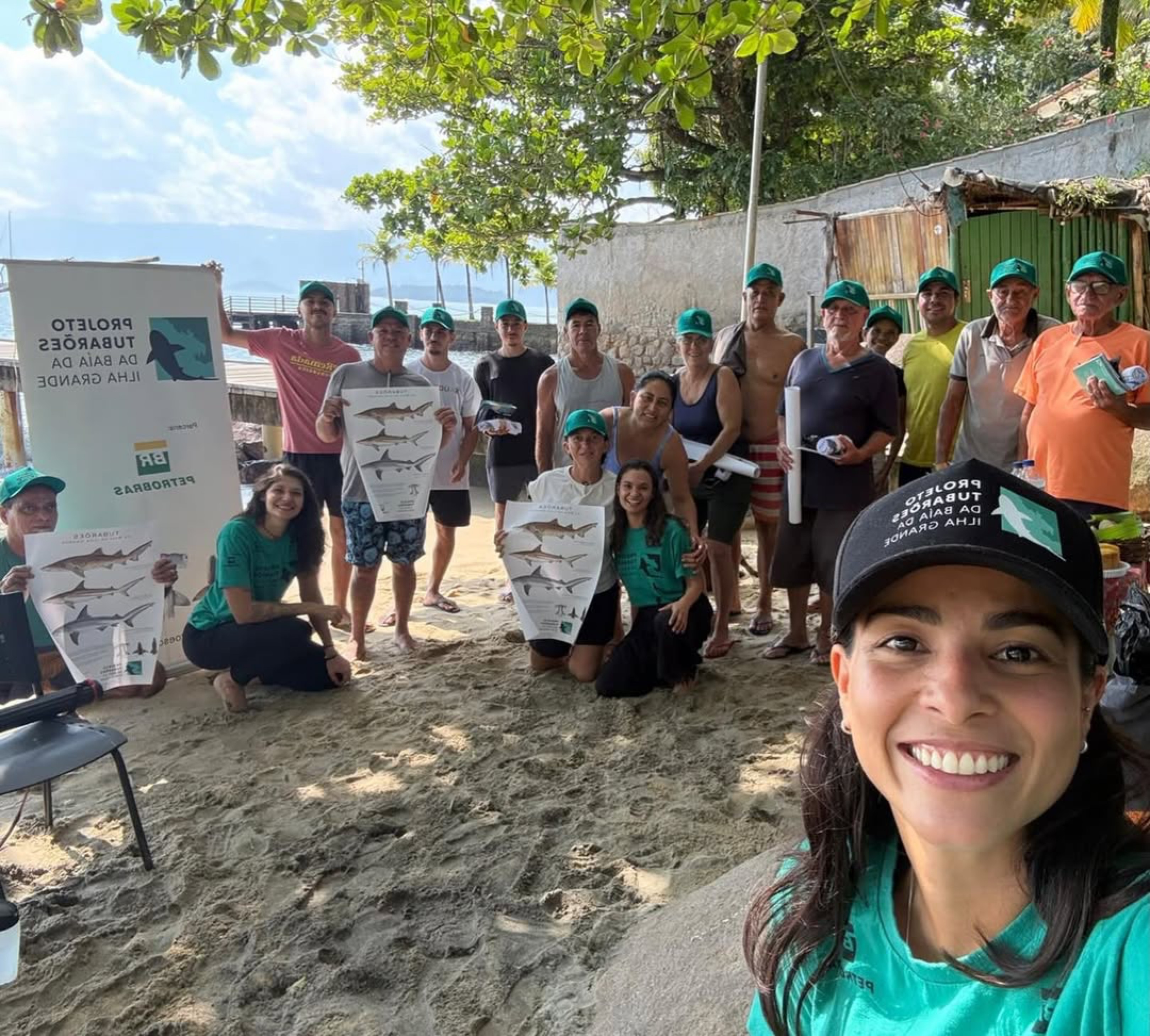 Mulher sorridente em primeiro plano, tirando selfie com um grupo de cerca de 15 pessoas em uma praia. Todos usam bon&eacute;s verdes e alguns seguram cartazes com imagens de tubar&otilde;es. Ao fundo, um banner branco com texto invertido e o mar