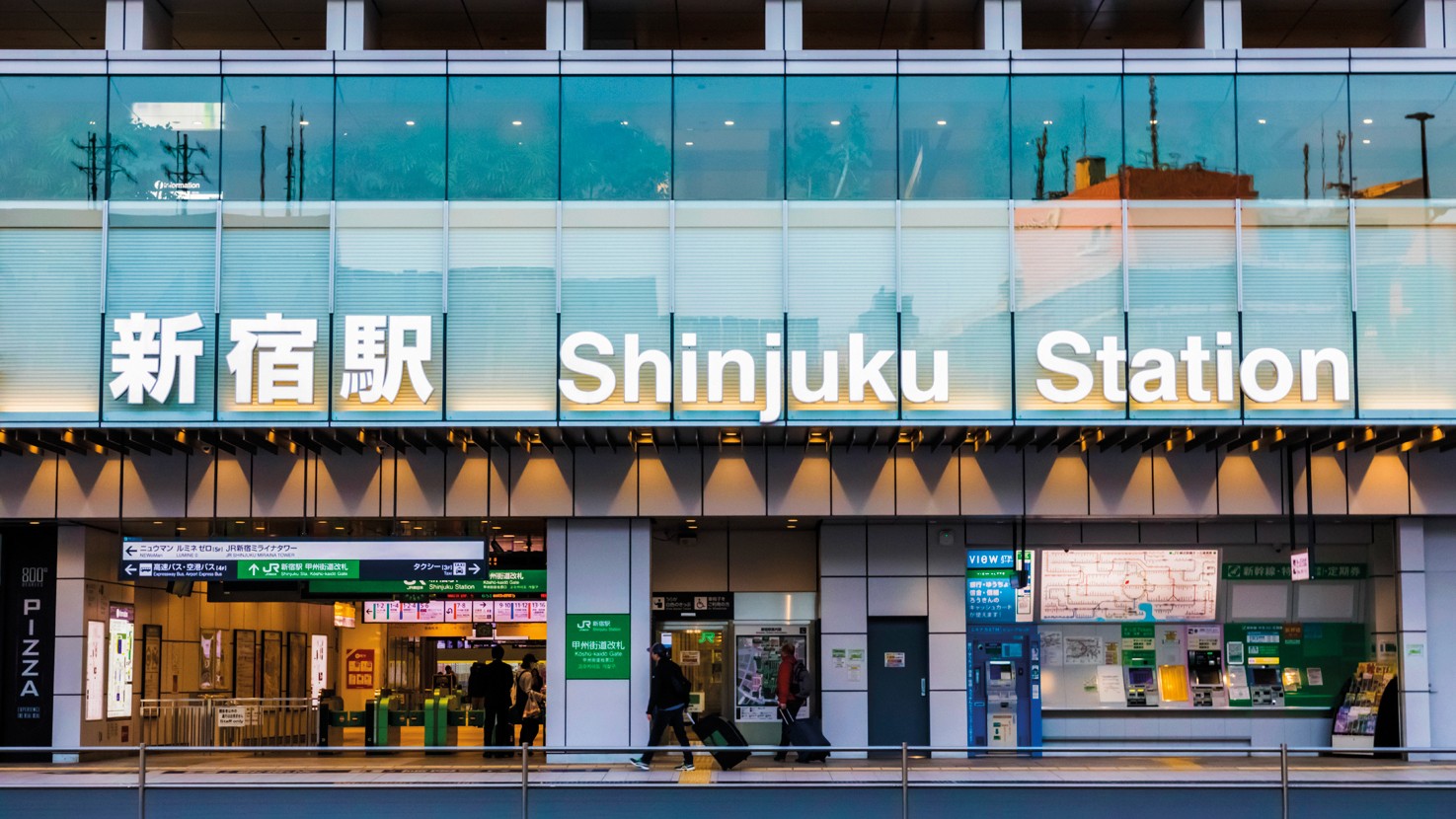 Fachada da Esta&ccedil;&atilde;o Shinjuku em T&oacute;quio, com o nome Shinjuku Station em letras brancas iluminadas e caracteres japoneses. Abaixo, pessoas entram e saem da esta&ccedil;&atilde;o, com placas de sinaliza&ccedil;&atilde;o e m&aacute;quinas de bilhetes vis&iacute;veis. A arquitetura moderna apresenta grandes janelas de vidro e detalhes em metal, refletindo o c&eacute;u azul claro.